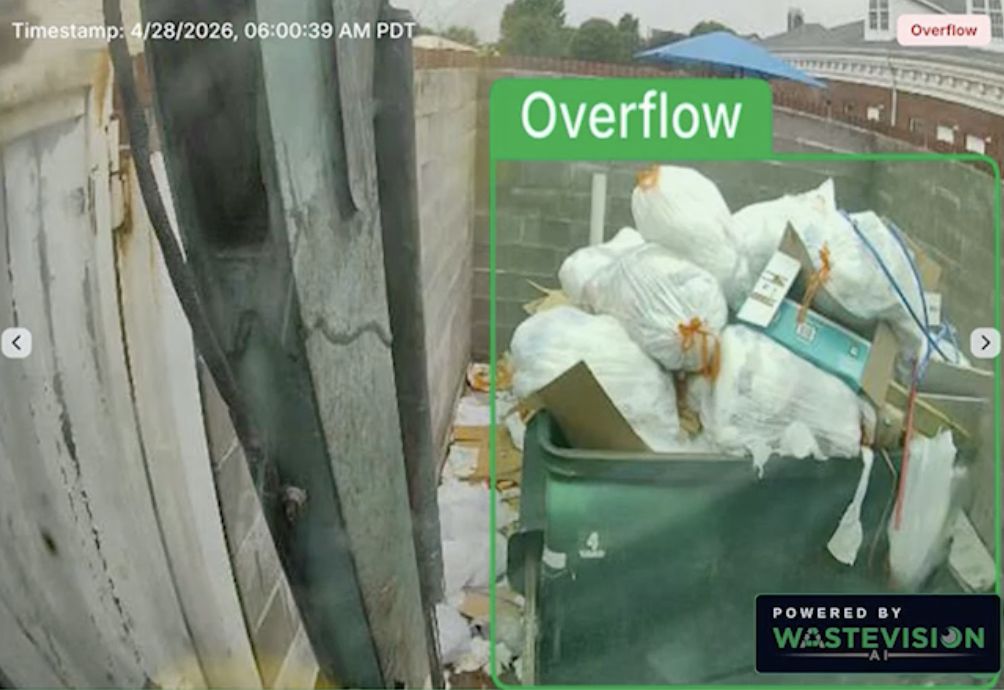 WasteVision AI camera frame of an overflowing green front-load dumpster piled with trash bags and cardboard, outlined by a green box labeled Overflow, with a timestamp in the corner